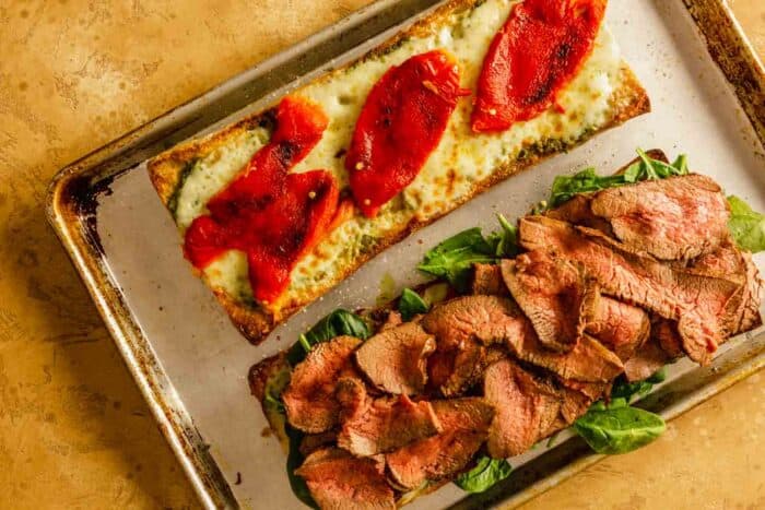 two halves of ciabatta bread on a baking sheet, one half topped with cheese and roasted bell peppers, the other half topped with spinach and sliced steak
