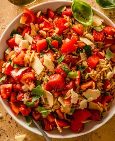 chunks of tomatoes and watermelon tossed with orzo pesto and basil in a large white bowl