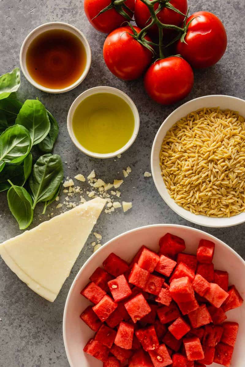 diced watermelon, vine-ripe tomatoes, olive oil, vinegar, basil, dry orzo pasta, and chunk of pecorino cheese on a counter