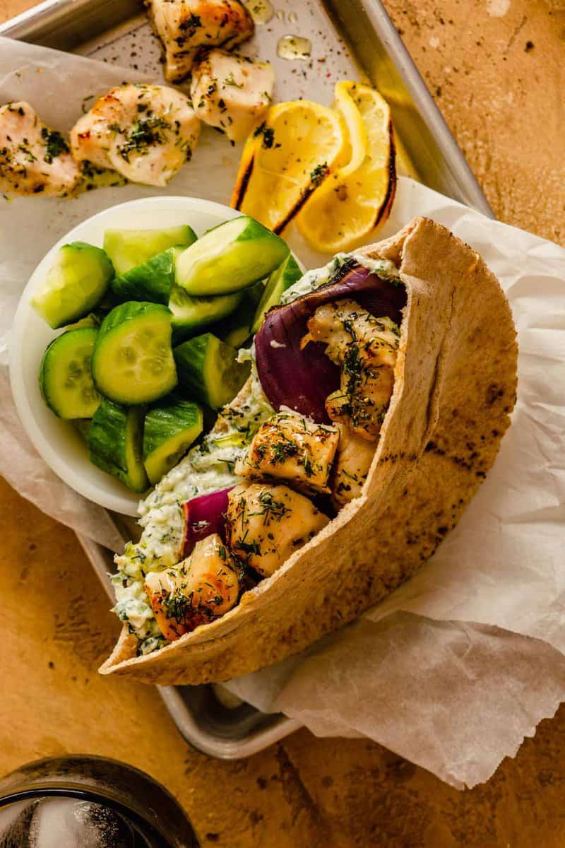 chunks of herb-coated chicken in a pita bread with tzatziki and a bowl of cucumbers on the side