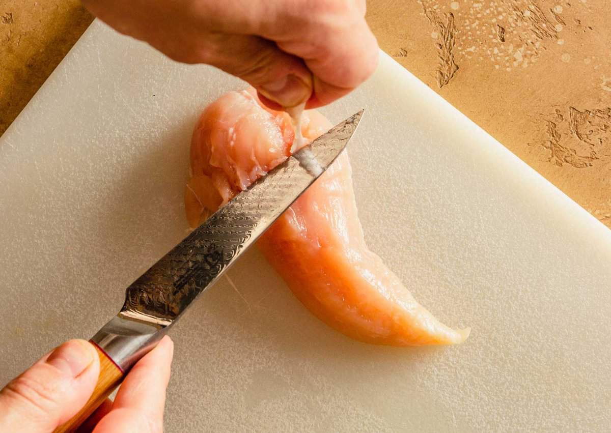 a set of hands cutting a tendon out of a chicken tender on a white plastic cutting board