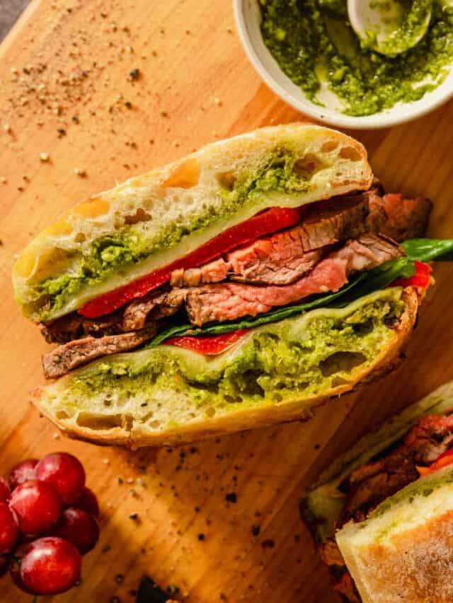 steak sandwich on a wood cutting board with pesto and grapes set around it