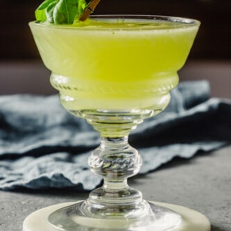 green-colored cocktail in a coupe glass set on a white coaster with a basil leaf as a garnish