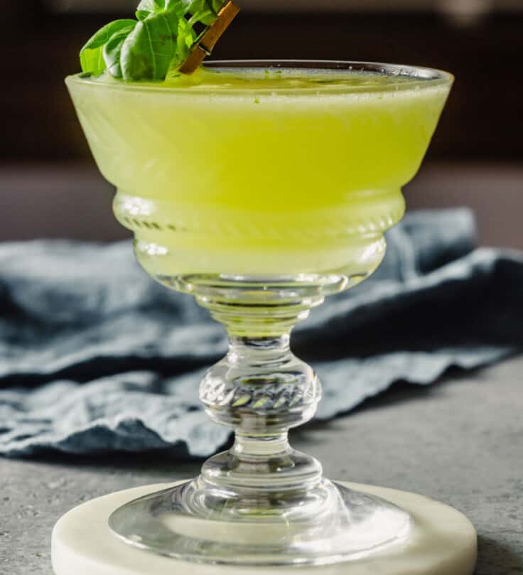 green-colored cocktail in a coupe glass set on a white coaster with a basil leaf as a garnish