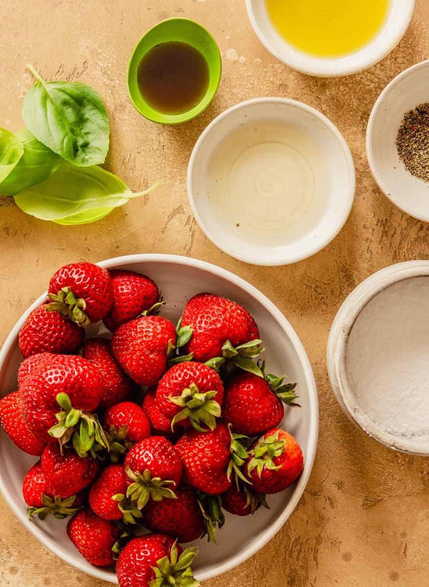 strawberries, olive oil, vinegar, basil and maple syrup set out on a counter in white bowls