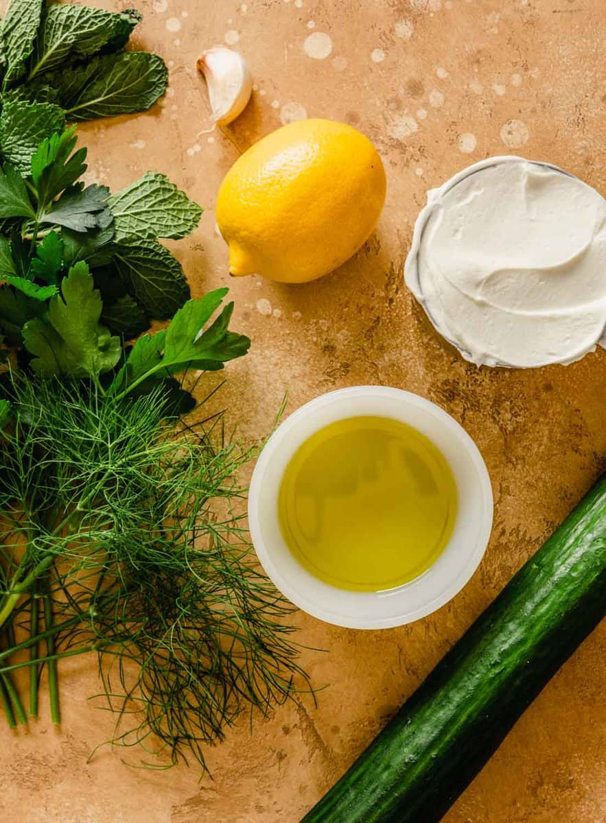 cucumber, yogurt, lemon, olive oil, garlic clove, and fresh herbs arranged on a counter