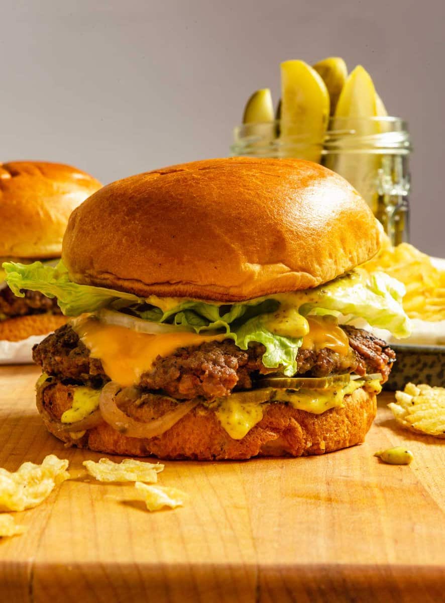 smash burger on a wooden cutting board with pickles set in the background