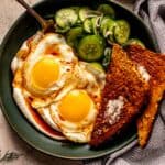 fried eggs placed over yogurt mixture in a matte green bowl topped with a cucumber and herb salad, chili oil and toast