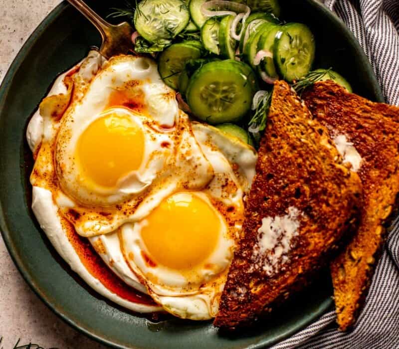 fried eggs placed over yogurt mixture in a matte green bowl topped with a cucumber and herb salad, chili oil and toast