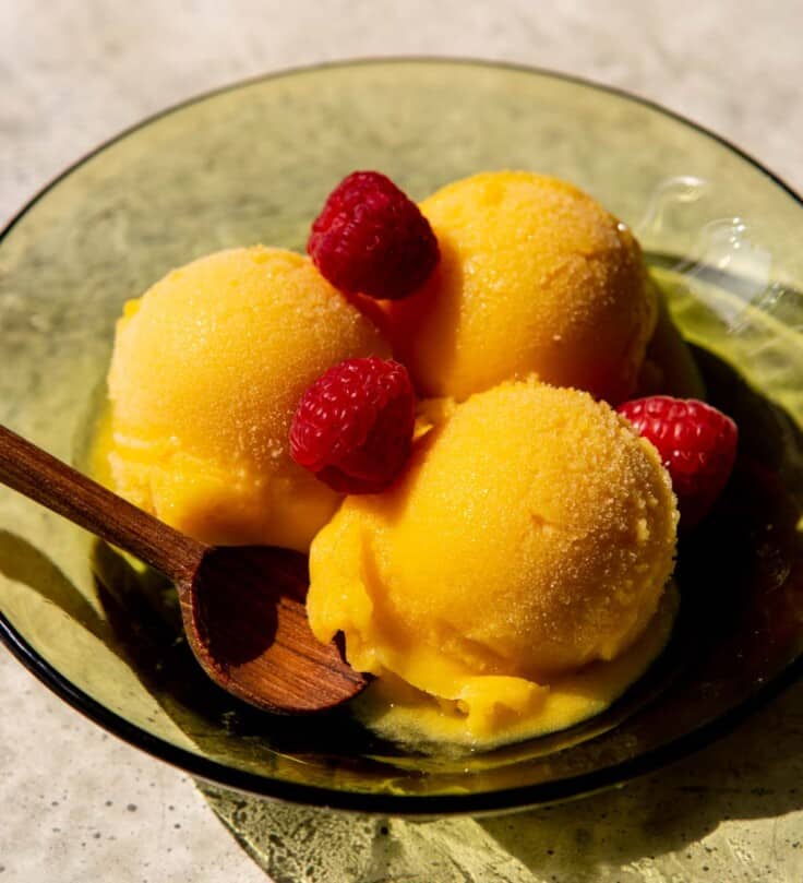 scoops of mango sorbet in a green glass bowl topped with fresh raspberries