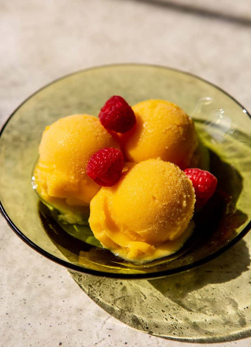 scoops of mango sorbet in a green glass bowl topped with fresh raspberries