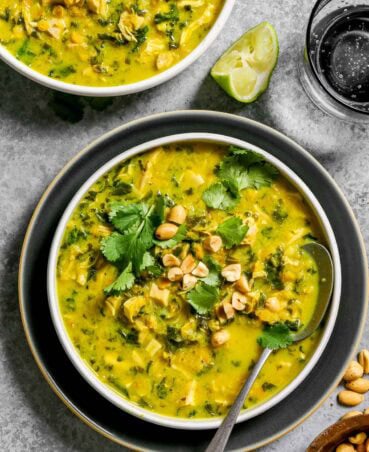 Chicken lentil soup in shallow white bowls, one bowl set on a blue plate with fresh cilantro and roasted peanuts sprinkled over top. Silver spoon set in soup bowl with a glass of bubbly water set to the side.