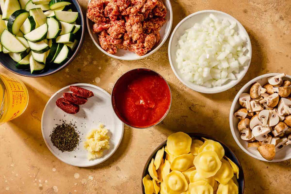spicy italian sausage, onion, ravioli, zucchini, garlic, tomato paste and oregano measured out and set out on a counter