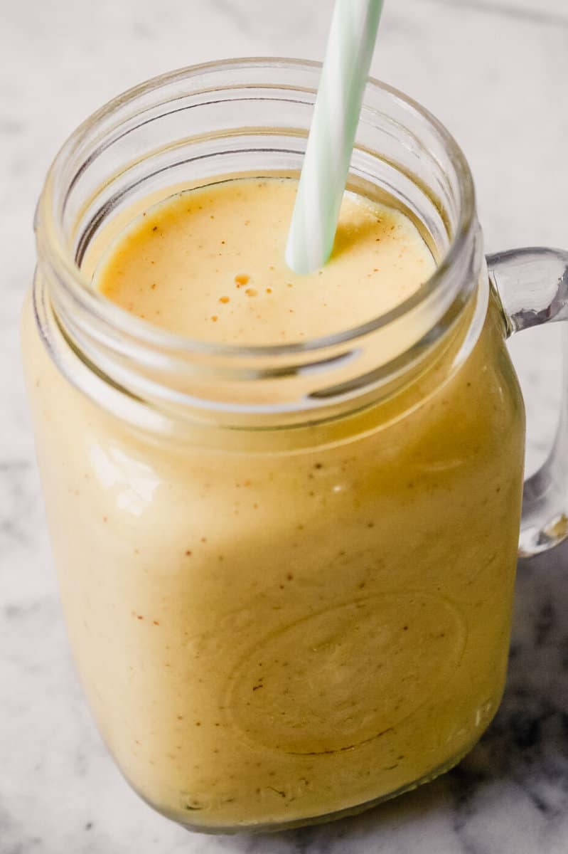 Side angle of a glass ball jar filled with a yellow-range smoothie set on a white marble table.