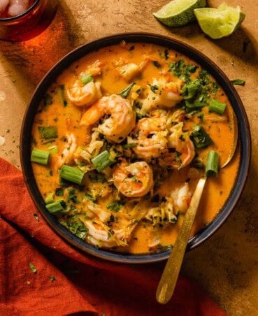 Thai shrimp soup with full pieces of shrimp, herbs, strands of cabbage and a red coconut curry broth in a shallow blue bowl and gold spoon.