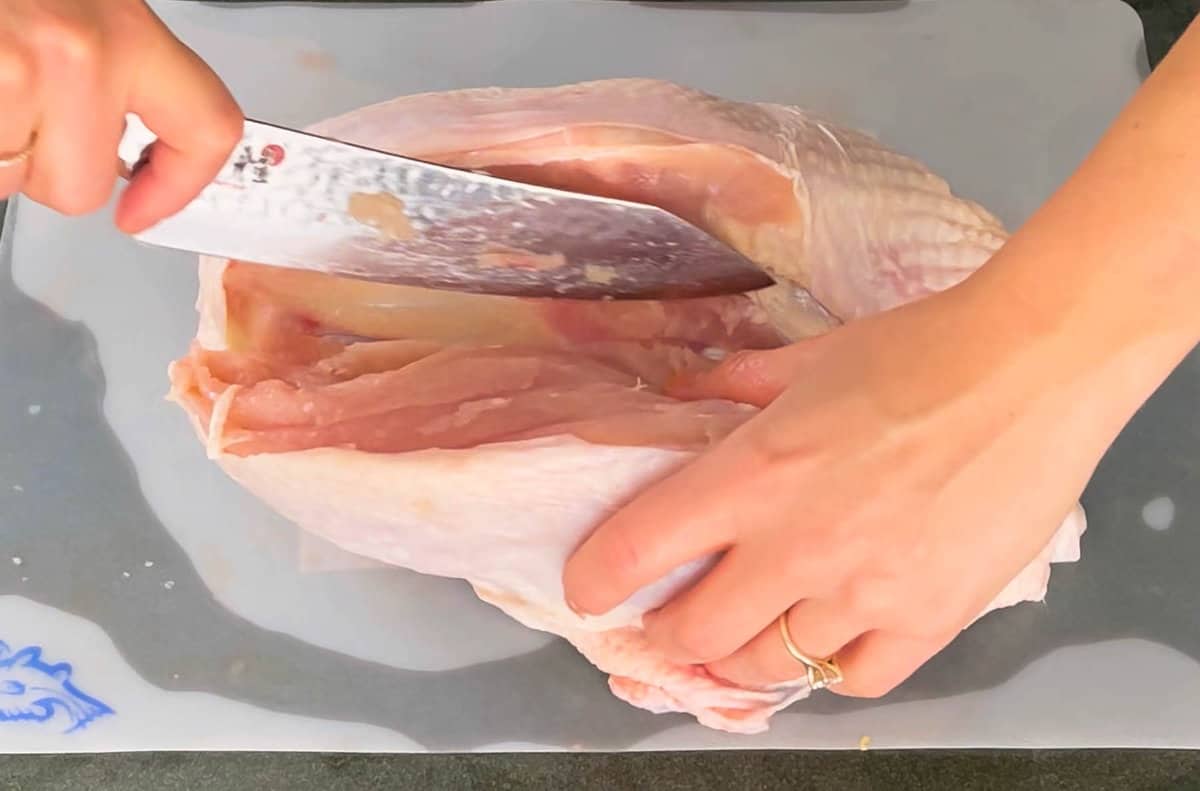 a set of hands breaking down a raw turkey with a chefs knife on a plastic cutting board