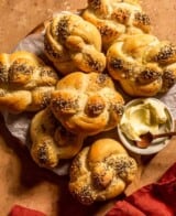 kaiser rolls stacked together on a wood board lined with parchment paper. a bowl of butter and a wooden spoon set next to the rolls