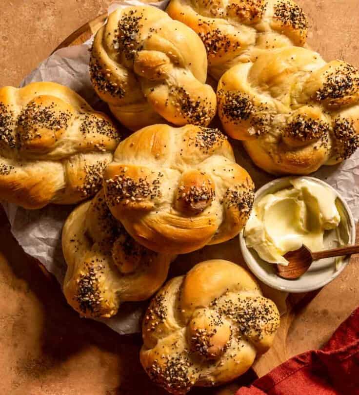 kaiser rolls stacked together on a wood board lined with parchment paper. a bowl of butter and a wooden spoon set next to the rolls