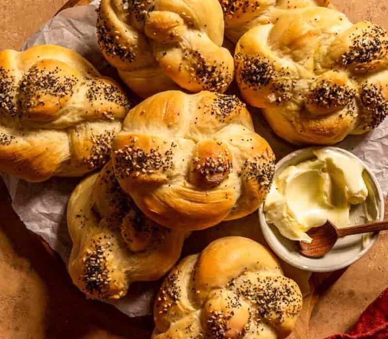 kaiser rolls stacked together on a wood board lined with parchment paper. a bowl of butter and a wooden spoon set next to the rolls