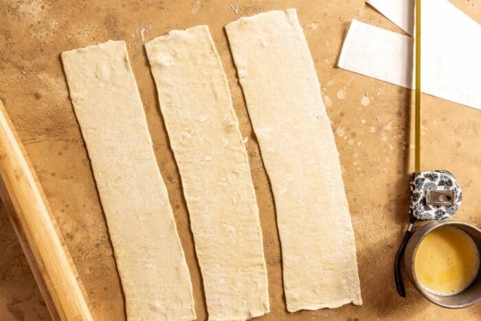 three strips of pastry dough on a counter with a measuring tape and a rolling pin