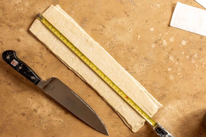 one strip of pastry dough on a counter with a measuring tape set on top and a chefs knife set nearby