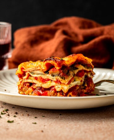 slice of lasagna on a white scalloped plate with a gold fork set on the plate and a glass of red wine set next to it