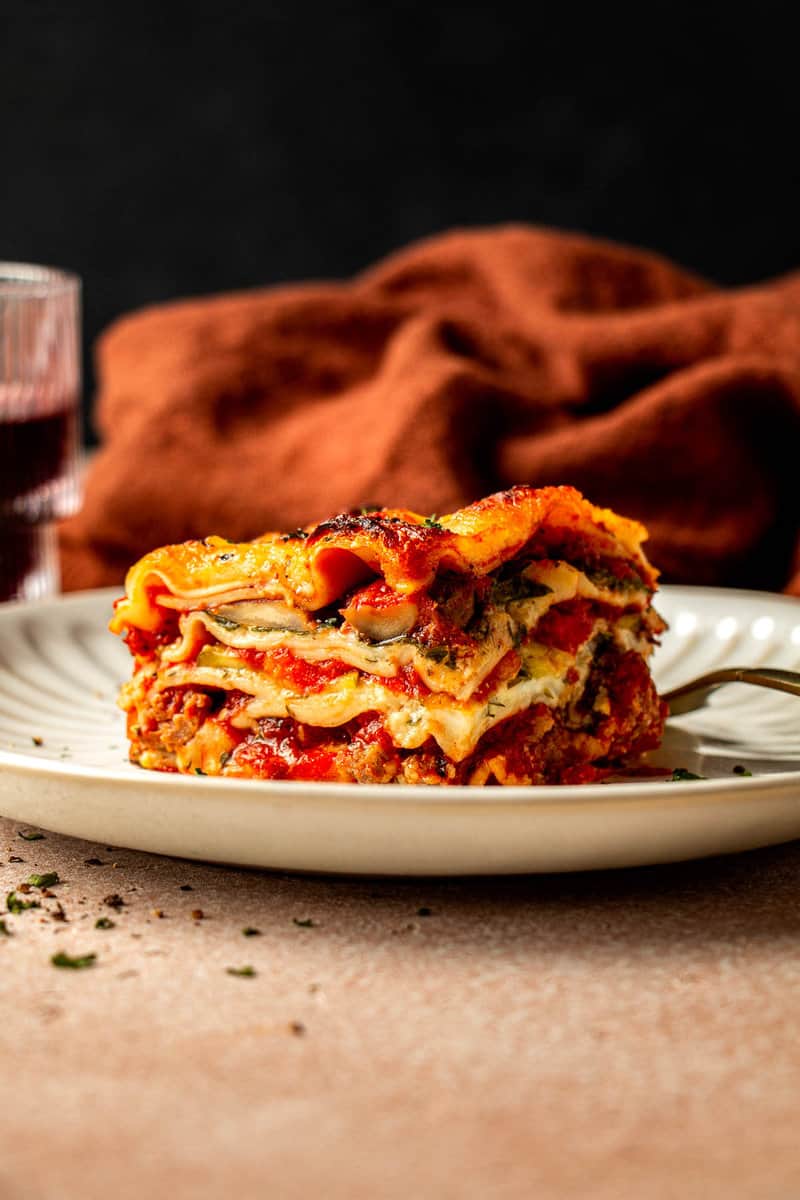 slice of lasagna on a white scalloped plate with a gold fork set on the plate and a glass of red wine set next to it