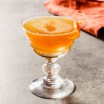 Orange-hued cocktail in a coupe glass with a burnt orange napkin set behind it