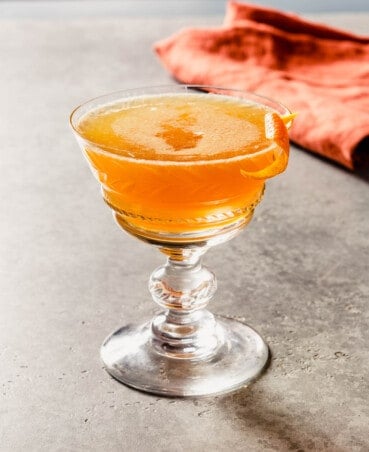 Orange-hued cocktail in a coupe glass with a burnt orange napkin set behind it