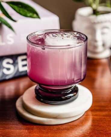 purple-hued cocktail in a short rocks glass set on white coasters on a wood table with plants behind it