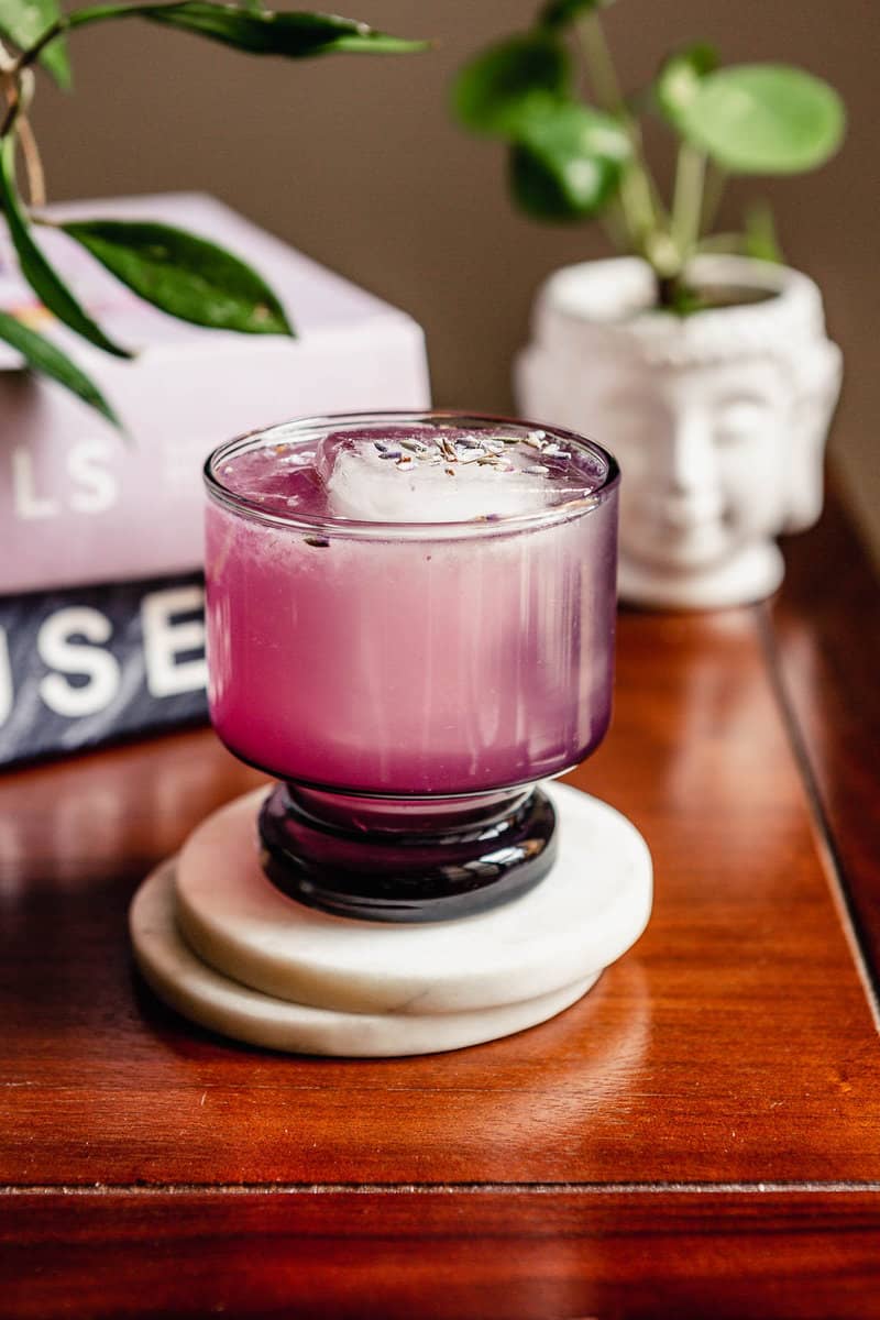 purple-hued cocktail in a short rocks glass set on white coasters on a wood table with plants behind it