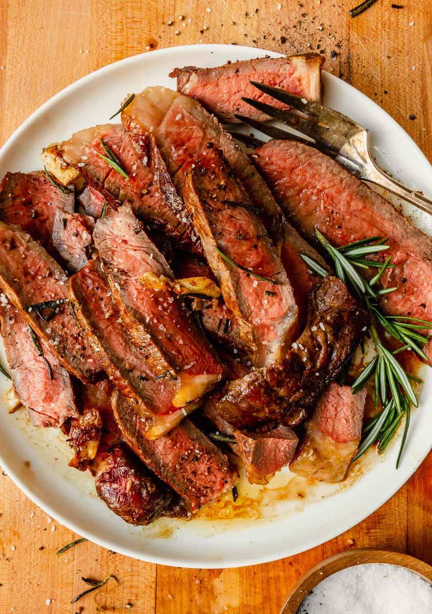 sliced rare steak on a white plate set on a wood cutting board with rosemary scattered around it