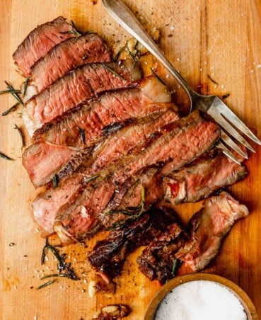 sliced rare steak on a wood cutting board with rosemary scattered around it