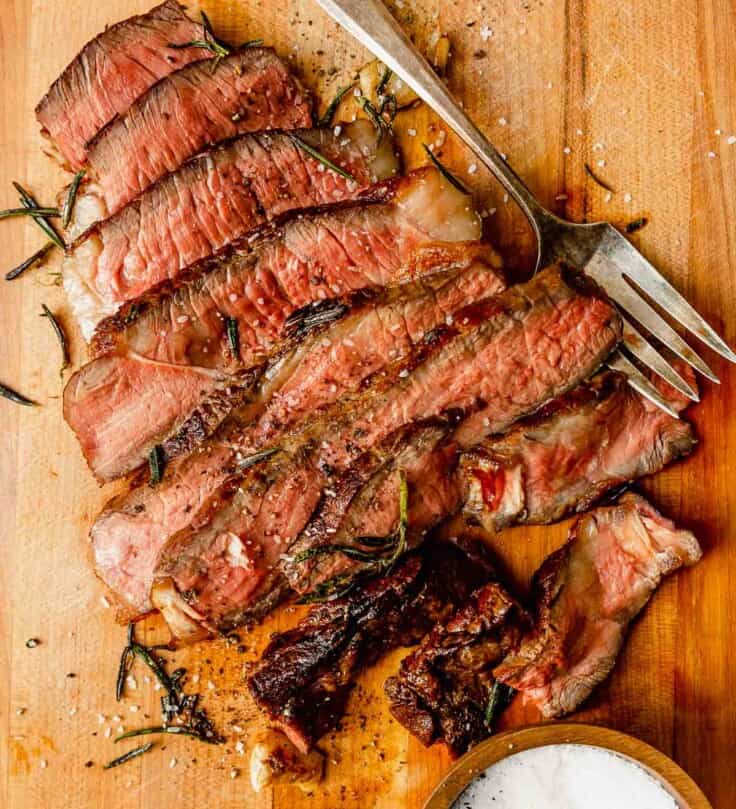 sliced rare steak on a wood cutting board with rosemary scattered around it