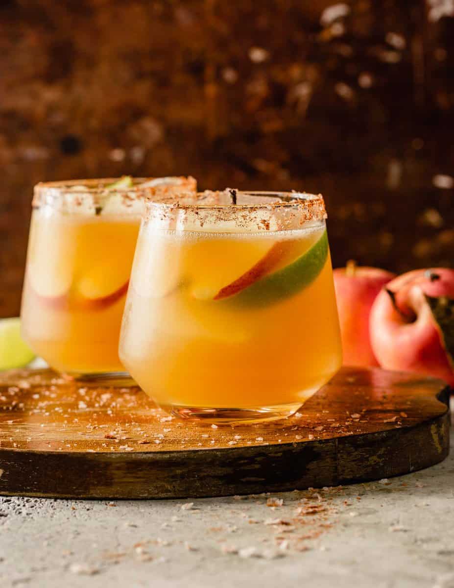 orange-hued cocktail in a rocks glass with ice and garnished with apple slices. Glasses set on a wood cutting board.