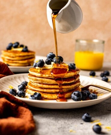 stack of pancakes on a white plate with a fork. pancakes topped with blueberry, lemon zest, and maple syrup