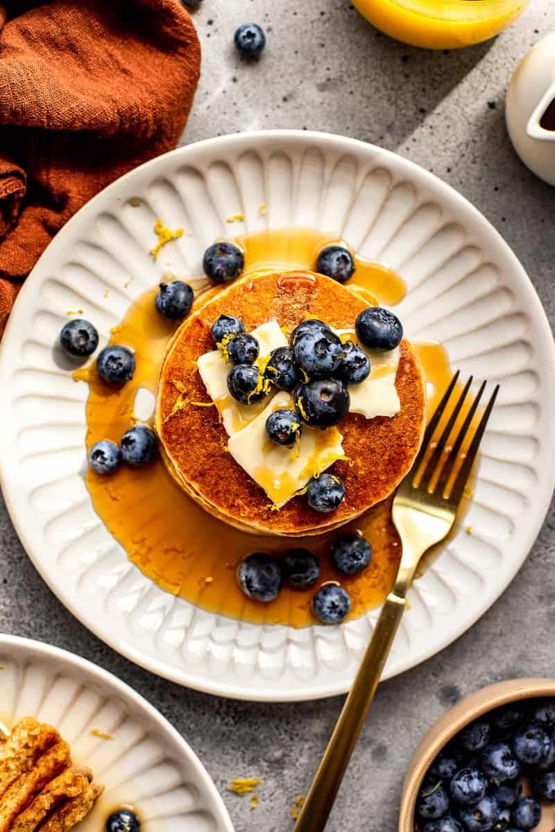 pancakes on a white plate topped with blueberries, butter and maple syrup