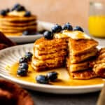 stack of pancakes on a white plate with a fork. pancakes topped with blueberry, lemon zest, and maple syrup