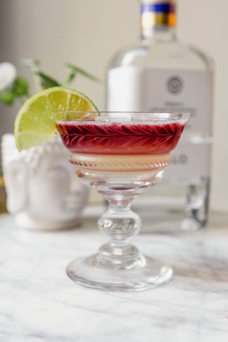 layered cocktail in a coupe glass set on a marble platter with a bottle of tequila and a plant set in the background