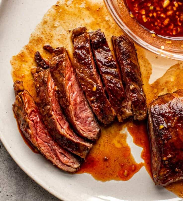 sliced steak on a plate with red sauce brushed over steak and in a bowl set next to steak