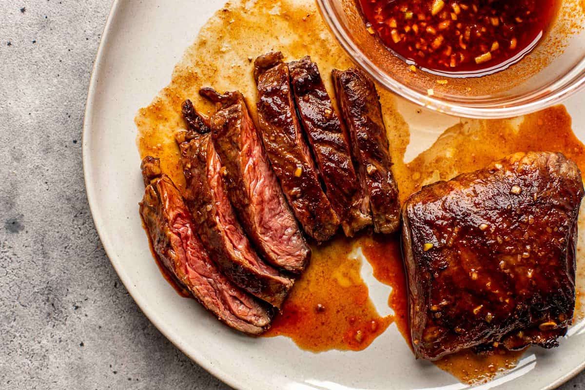 sliced steak on a plate with red sauce brushed over steak and in a bowl set next to steak