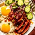 sliced steak, fried eggs, and cucumbers on a plate with orange napkins and glassware set around it