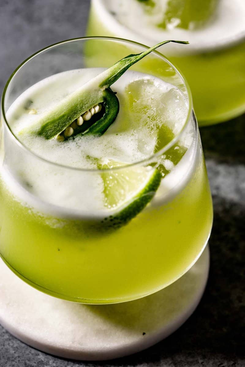 green cocktail with a lime wedge and jalapeno slice set on top. Cocktail set on a white coaster