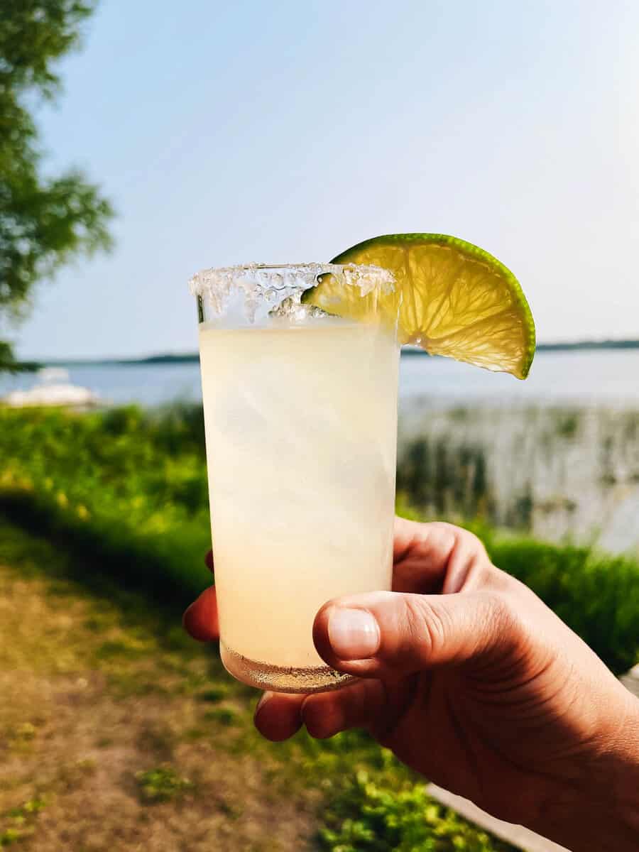 someone holding a margarita in a tall glass with a lime wedge on the rim with a lake in the background