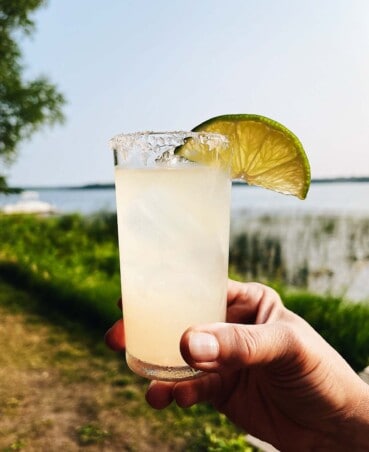 someone holding a margarita in a tall glass with a lime wedge on the rim with a lake in the background