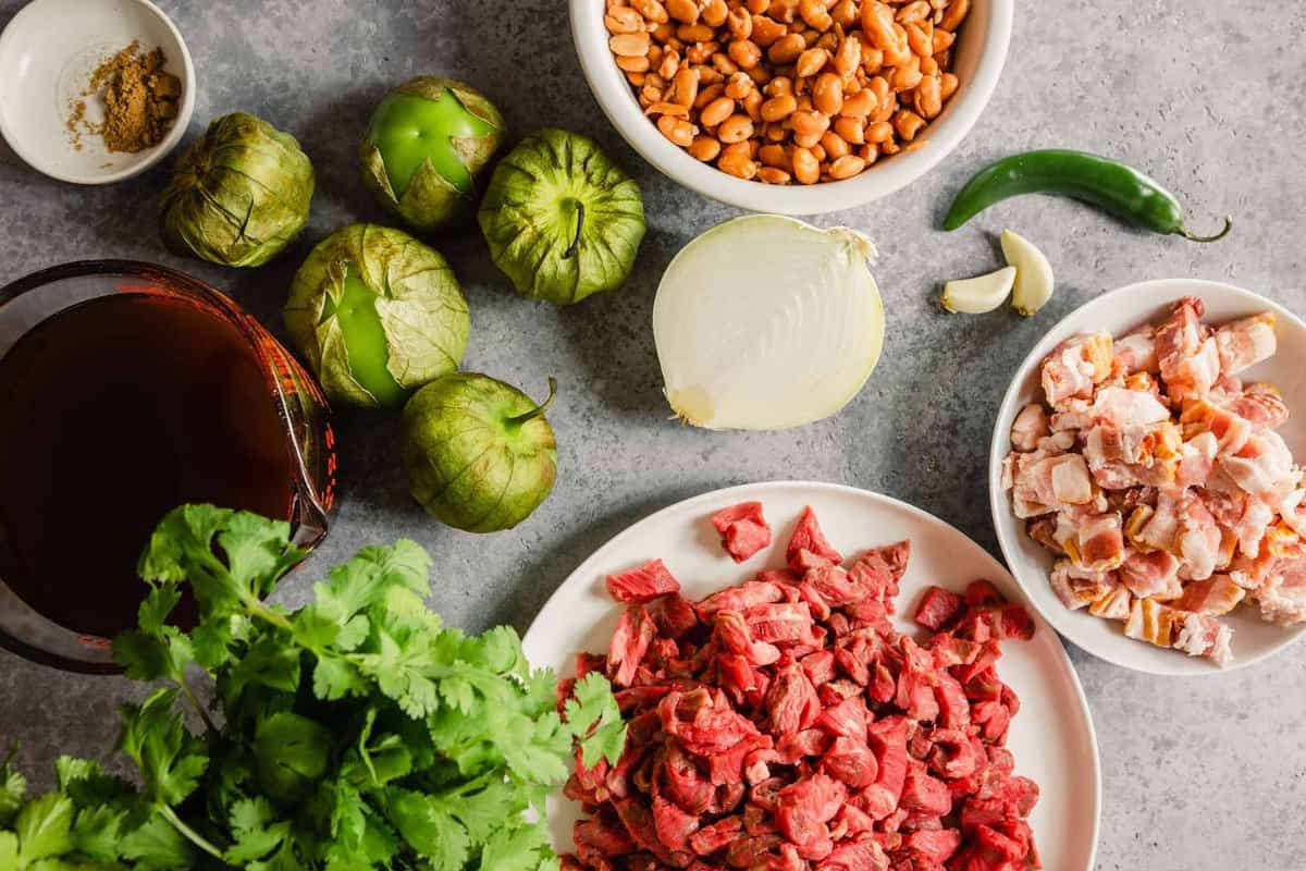 diced beef, diced bacon, cilantro, tomatillos, beans, onion, serrano, and garlic measured out on a counter