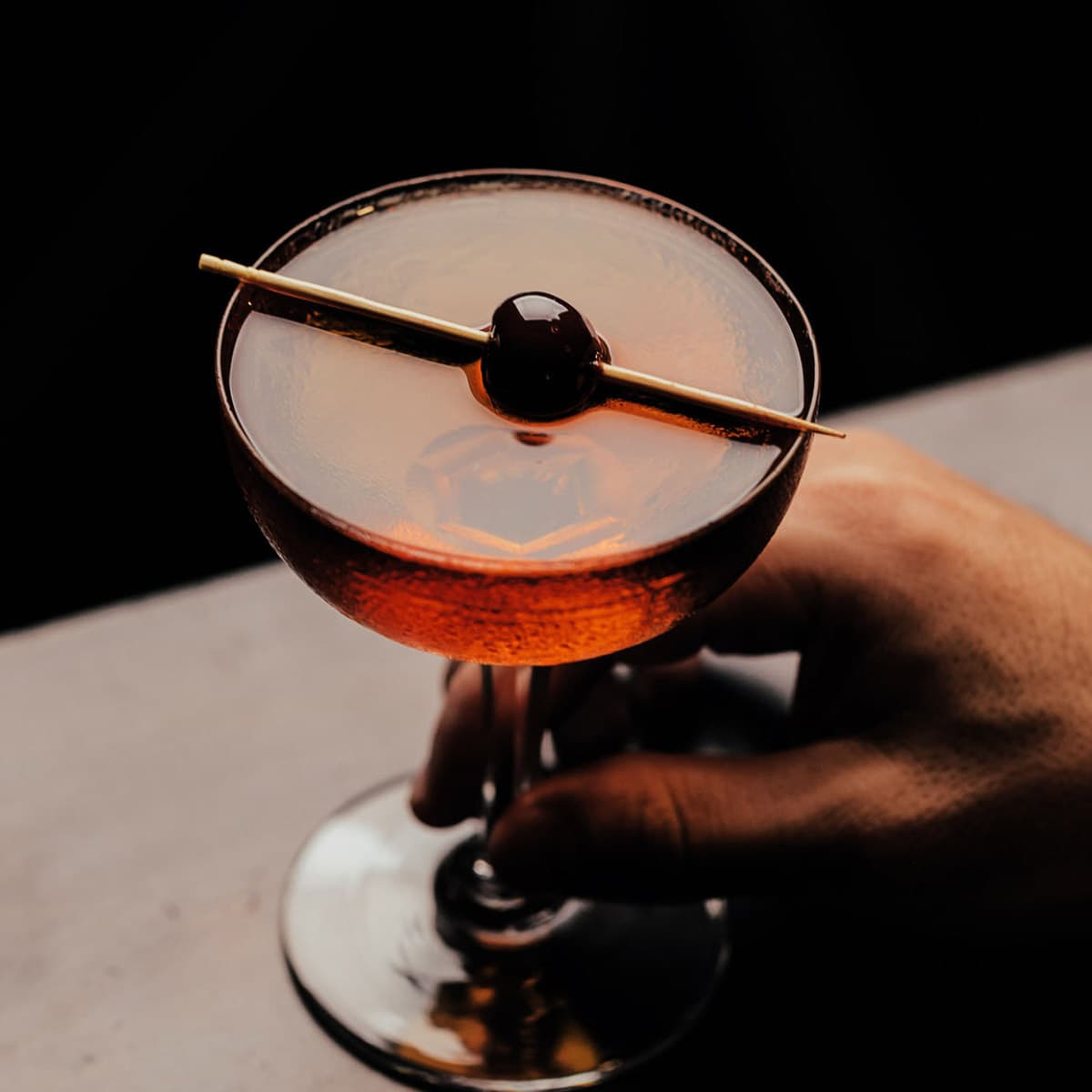 manhattan cocktail in a coupe glass garnished with a cherry. someone's hand holding the cocktail