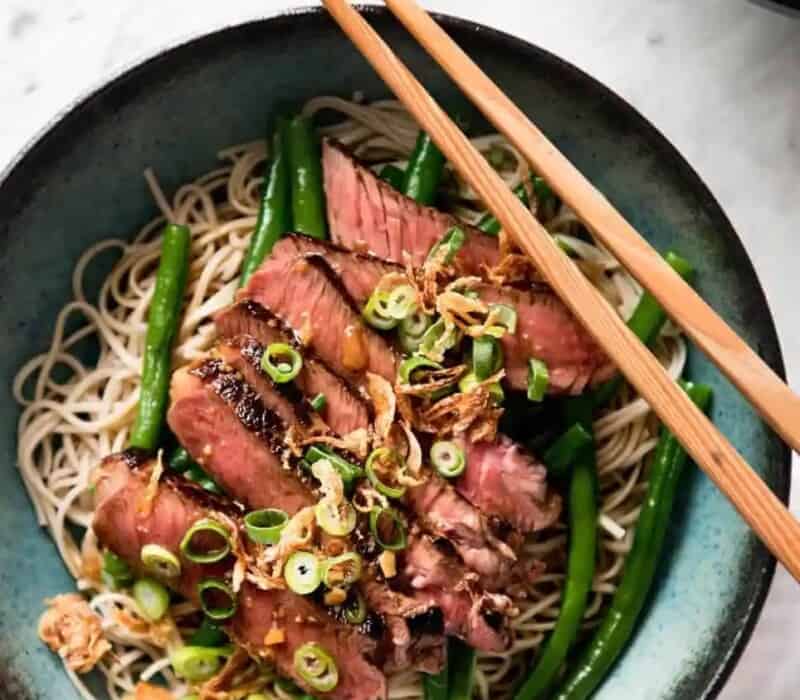 sliced steak set atop soba noodles in a teal bowl, topped with scallions. chopsticks set on top of the bowl