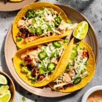 light pink plate with three salmon tacos set on it with sauce, water and limes set around it