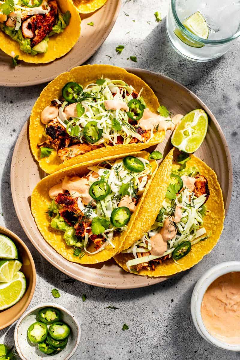 light pink plate with three salmon tacos set on it with sauce, water and limes set around it